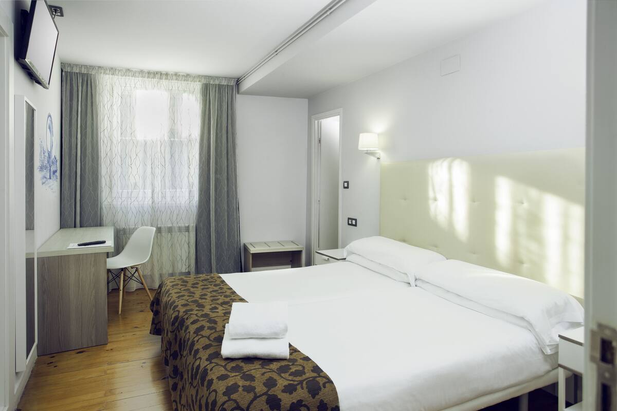 Une chambre d’hôtel comprenant un lit, un bureau, une chaise, une table de chevet, une lampe et une fenêtre avec des rideaux.