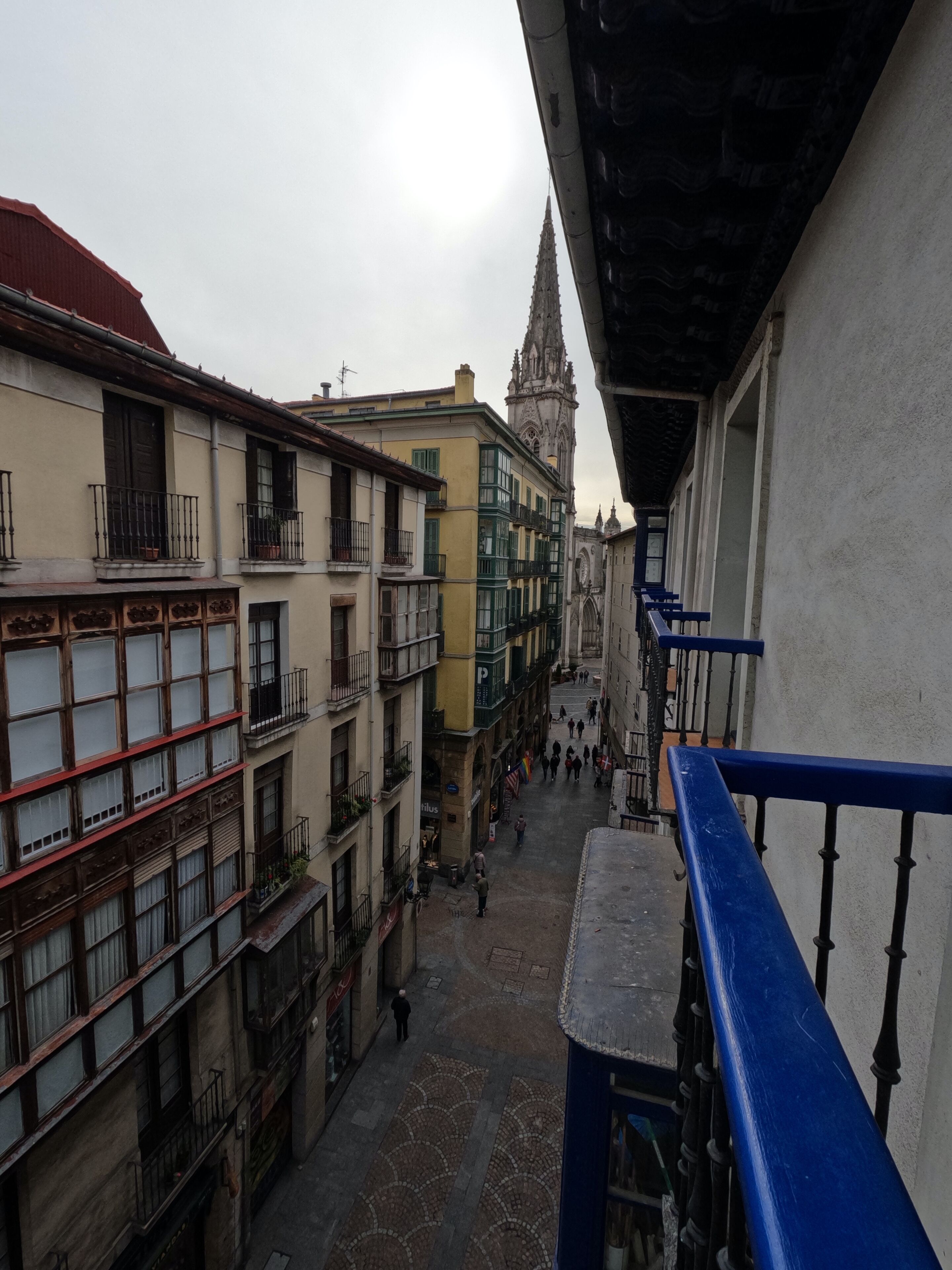 Una calle estrecha flanqueada por edificios, una barandilla azul y una torre al fondo.