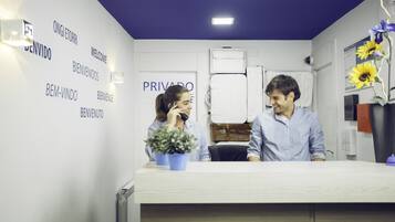 Una zona d'entrada amb un taulell de recepció blanc, una planta en test i una paret amb indicadors de benvinguda en diversos idiomes.