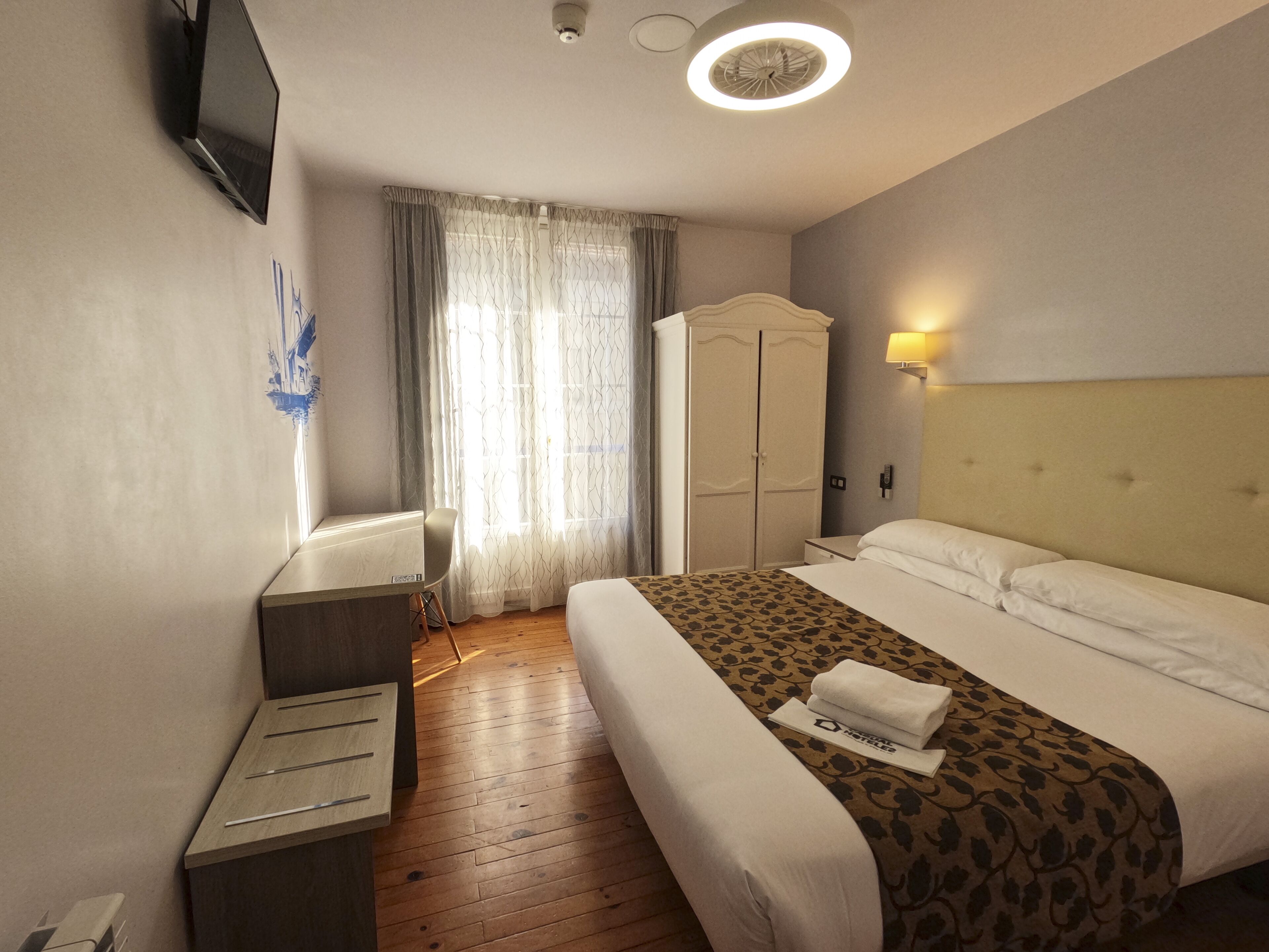 A hotel room with a bed, a desk, a wardrobe, and a TV mounted on the wall.