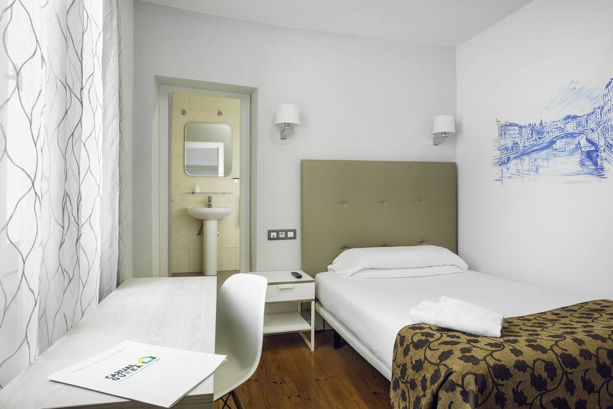 Une chambre d’hôtel comprenant un lit, un bureau, une chaise, une petite table, un miroir, une lampe fixée au mur et une œuvre d’art mural représentant un paysage urbain.