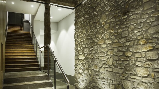 Una escalera con peldaños de madera y barandillas de metal que conduce a una habitación con pared de piedra e iluminación empotrada en el techo.