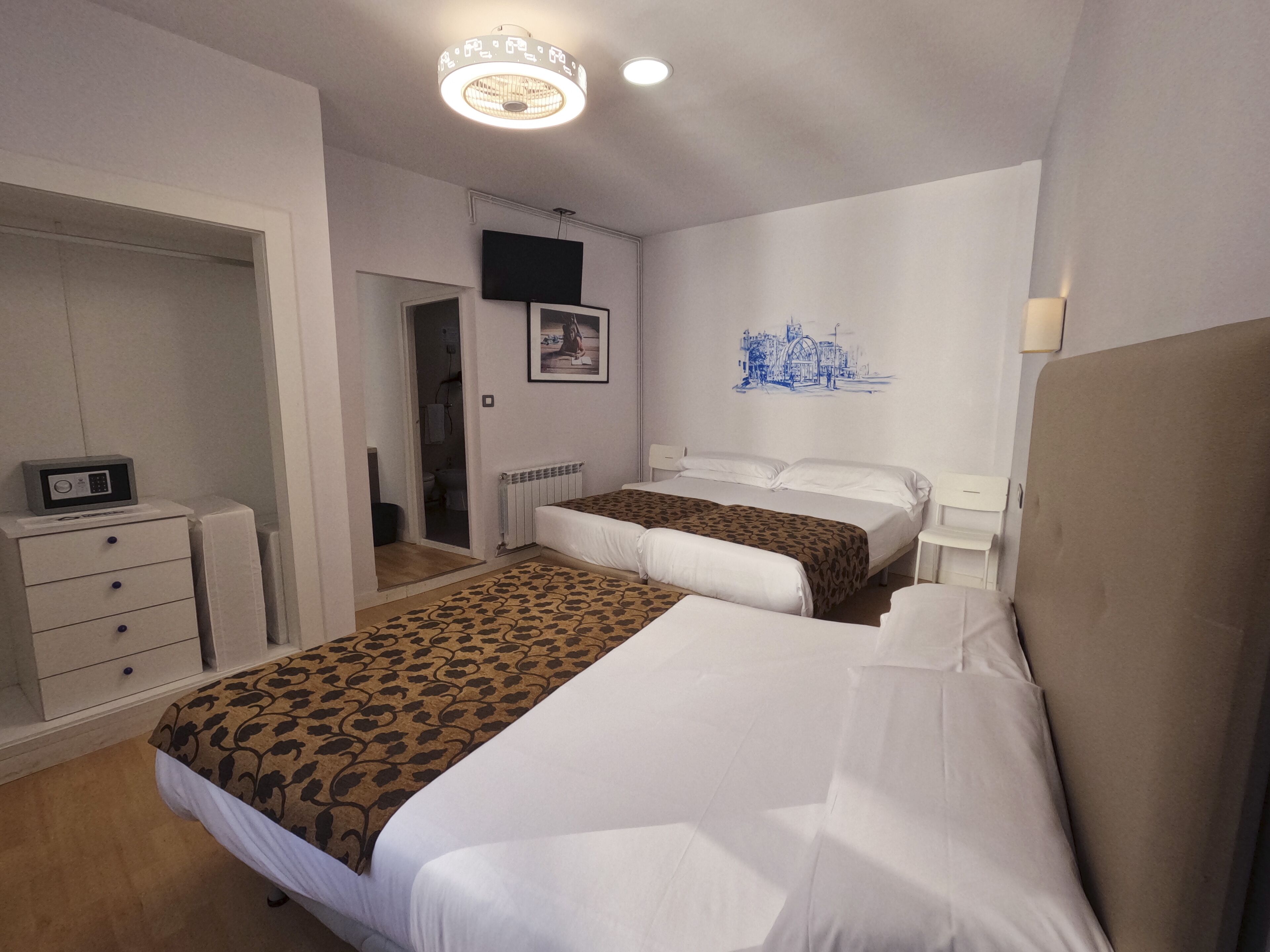 A hotel room with two beds, a wardrobe, a radiator, a television on the wall, and a cityscape artwork.