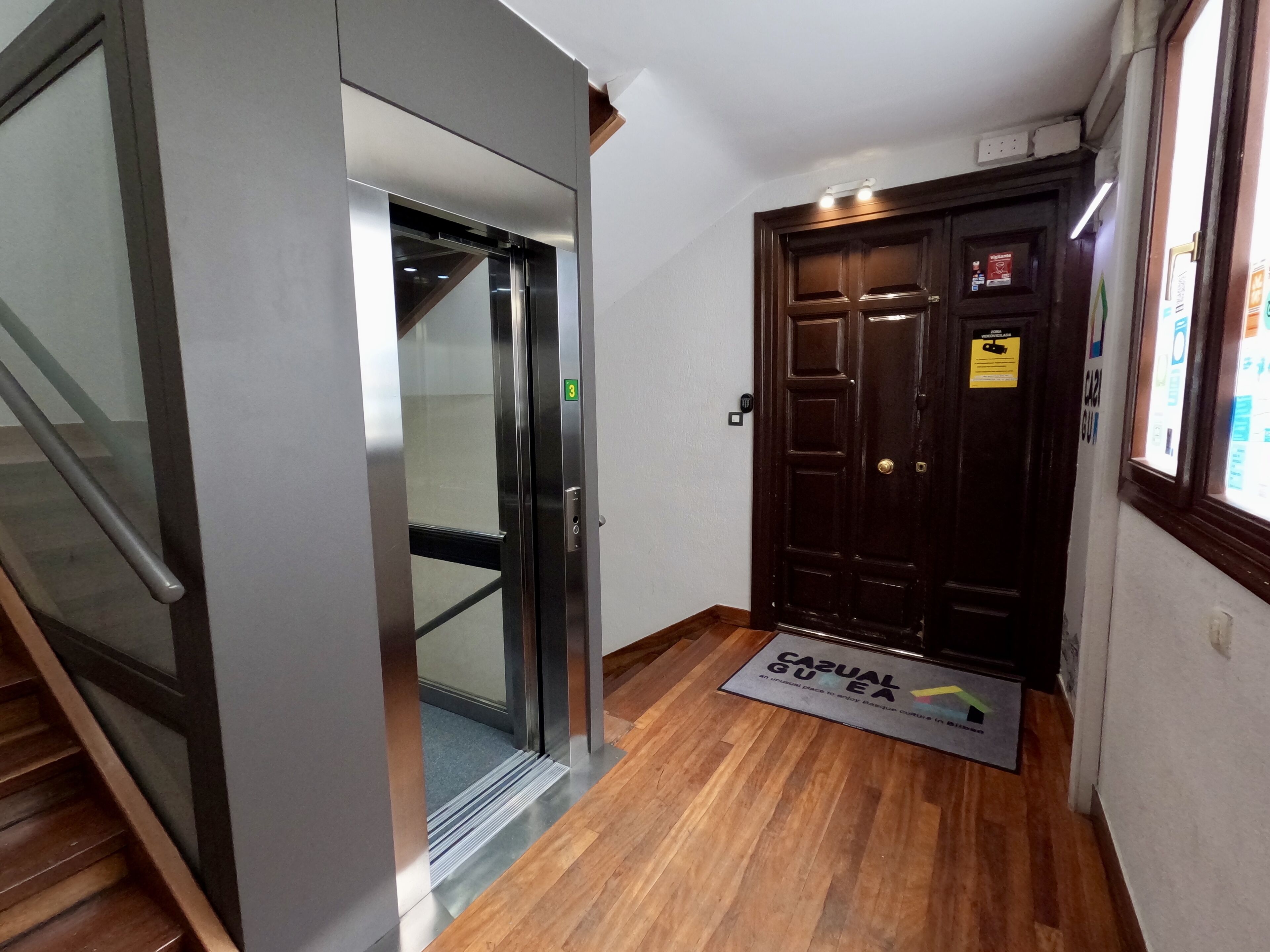 A hallway with a wooden floor, a glass elevator door, and a dark wooden door with a sign.