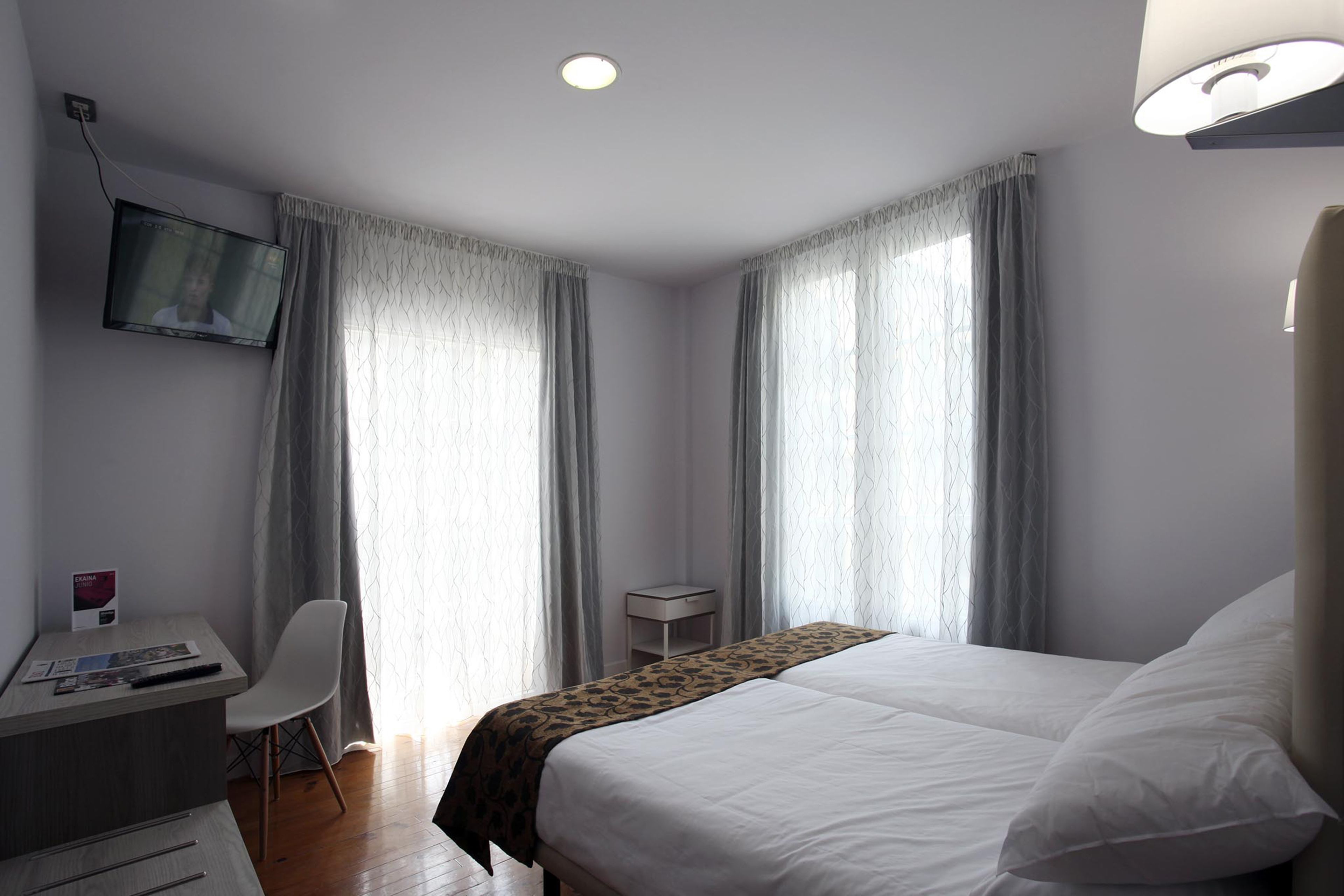 A hotel room with a bed, a desk with a chair, a TV mounted on the wall, and a small side table.