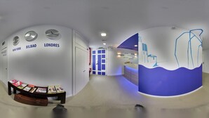 Un moderno vestíbulo de hotel con una pared curva que muestra relojes marcando la hora de Nueva York, Bilbao y Londres. Hay un mostrador de recepción con diseño azul y blanco, y un pequeño rincón de descanso con revistas.