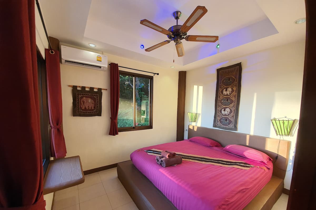 A bedroom with a bed, a ceiling fan, an air conditioner, a window with curtains, and a wall tapestry.