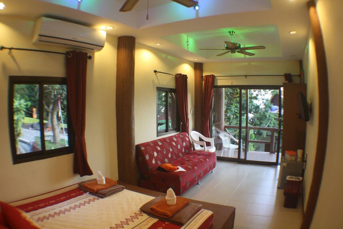 A hotel room with a bed, a red sofa, a balcony with chairs, and a ceiling fan.