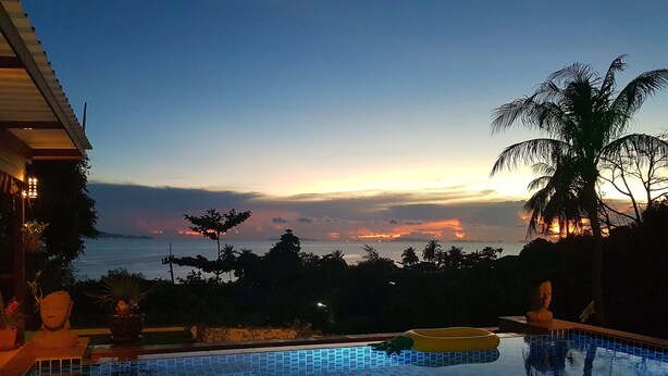A swimming pool with a view of the sunset over the ocean, surrounded by palm trees and a building with a tiled roof. - Moonstone Studio (Ko Pha-ngan)