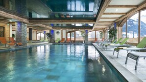An indoor swimming pool with multiple lounge chairs and tables, large windows offering mountain views, and a stone pillar with blue lighting. - CGH Résidences & Spas Les Chalets d'Angèle (Chatel)