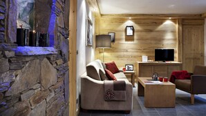 A cozy living room with a stone wall, wooden paneling, a sofa, a coffee table, a TV, and a fireplace. - CGH Résidences & Spas Les Chalets d'Angèle (Chatel)