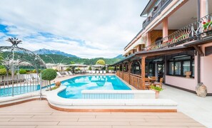 A swimming pool with a curved edge, surrounded by lounge chairs and a wooden deck. - Hotel Ariston (Levico Terme)