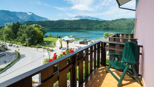 A balcony with a view of a lake, mountains, and a road with cars. - Hotel Ariston (Levico Terme)