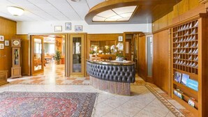 A lobby with a reception desk, a patterned rug, wooden paneling, and a bookshelf. - Hotel Ariston (Levico Terme)