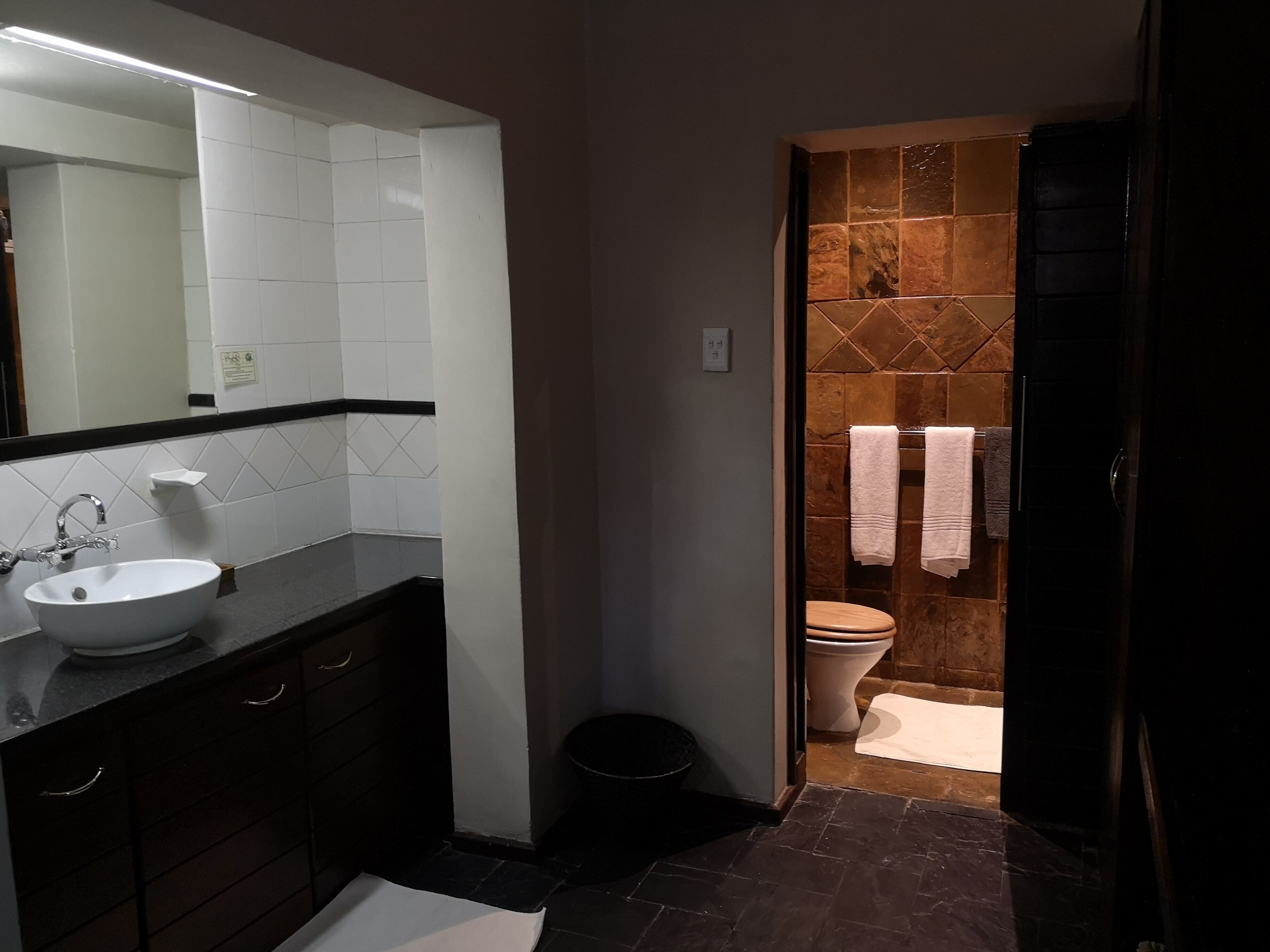 Une salle de bain avec un lavabo blanc, des meubles sombres, un miroir et un espace douche aux murs carrelés et une baignoire noire.