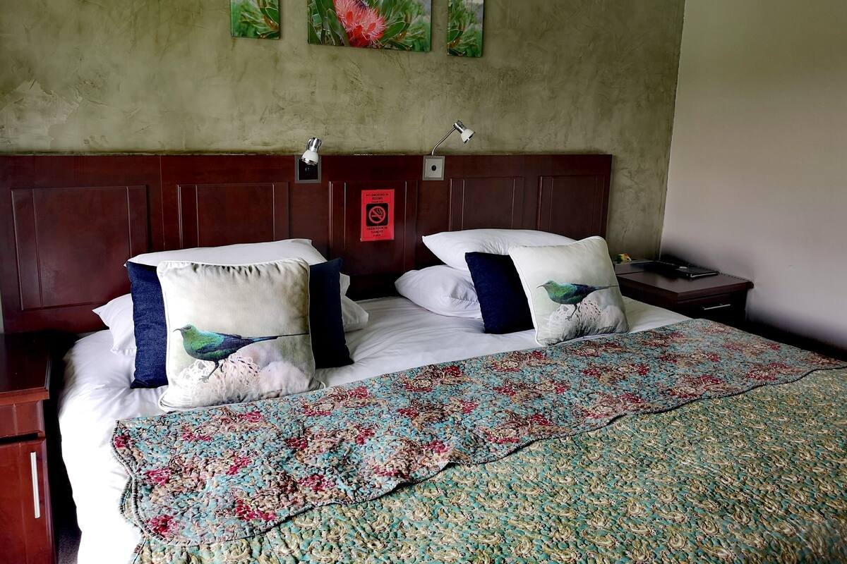 A hotel room with a wooden headboard, a bed with white linens, two pillows with bird designs, a floral-patterned bedspread, and three wall-mounted paintings of birds and flowers.