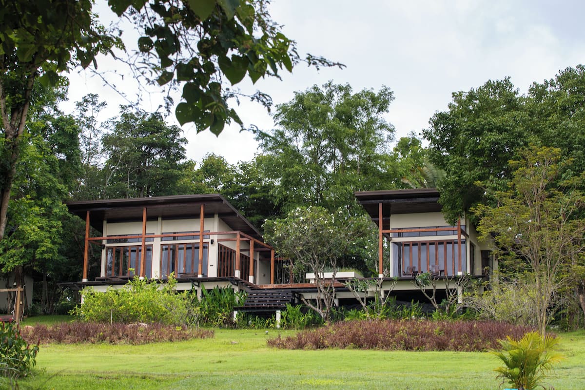 A modern two-story house with a large balcony, surrounded by lush greenery and a well-maintained lawn.