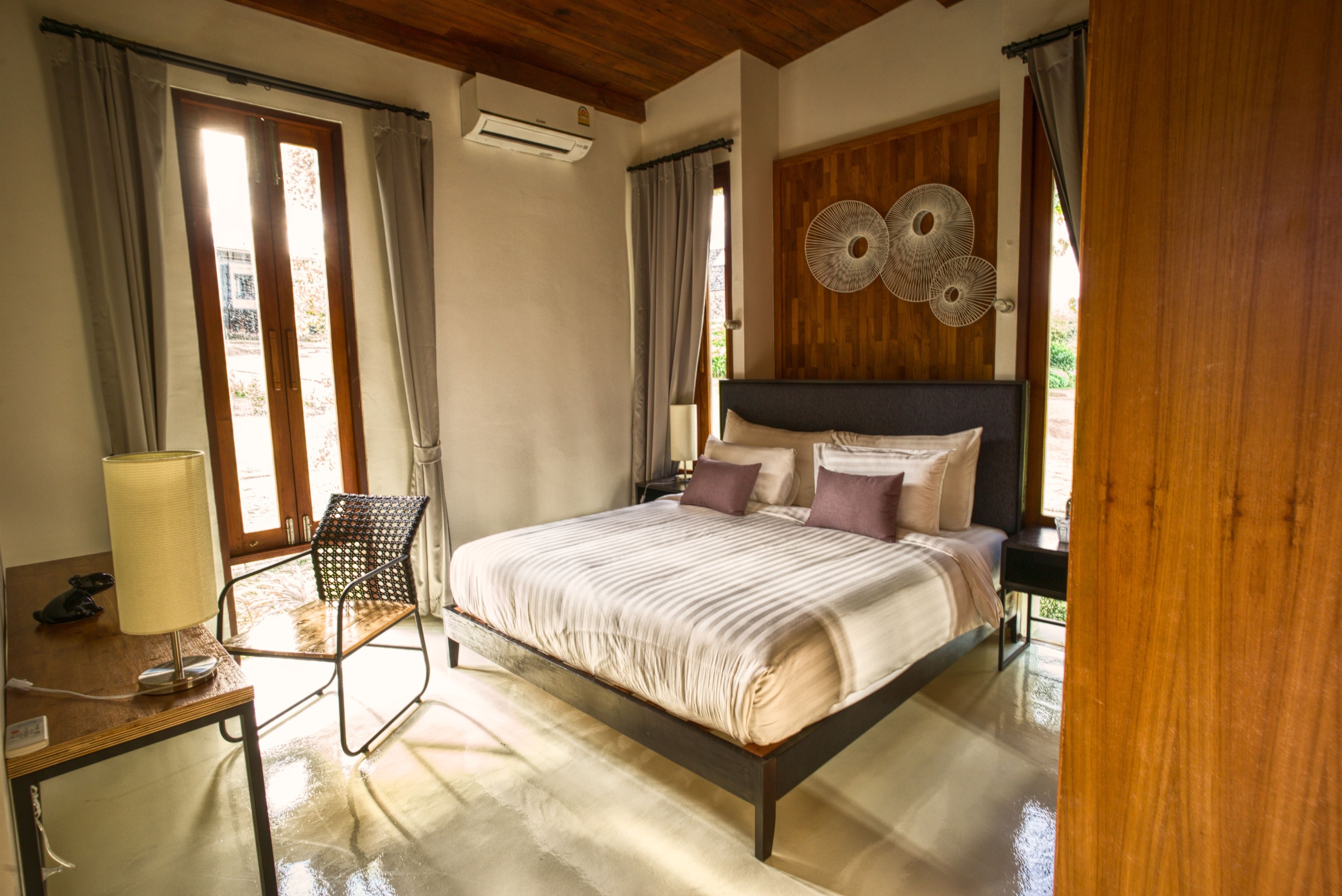 Un dormitorio con cama, una silla, una mesa con lámpara y un ventilador de pared.