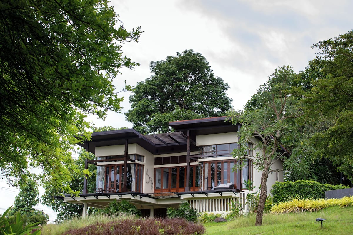 A two-story house with a balcony, surrounded by lush greenery and trees.