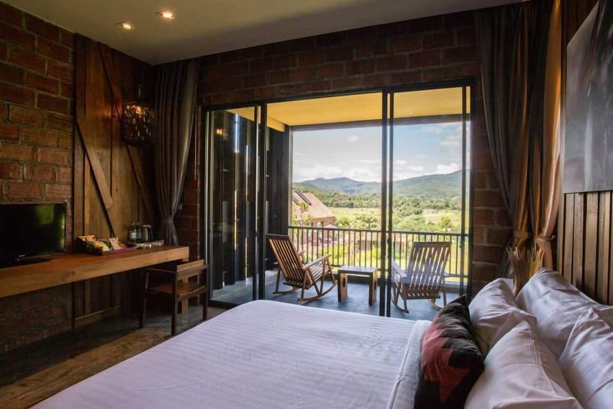 A bedroom with a large bed, a desk with a TV, and a balcony with chairs.