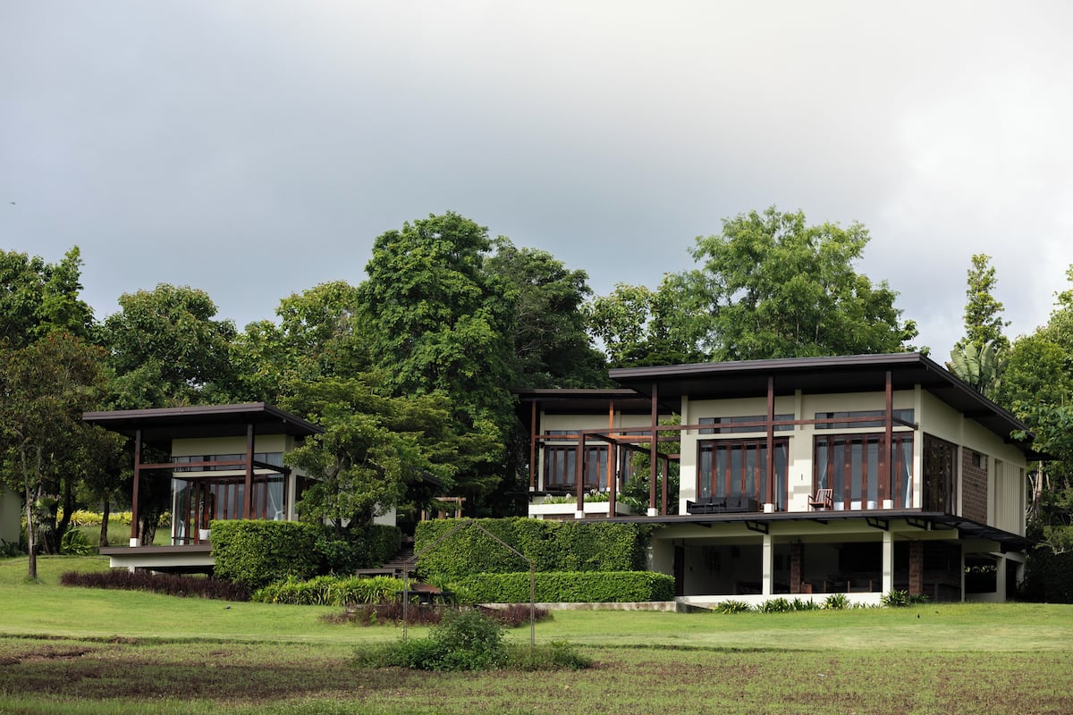 A modern two-story house with a balcony, surrounded by lush greenery and trees.