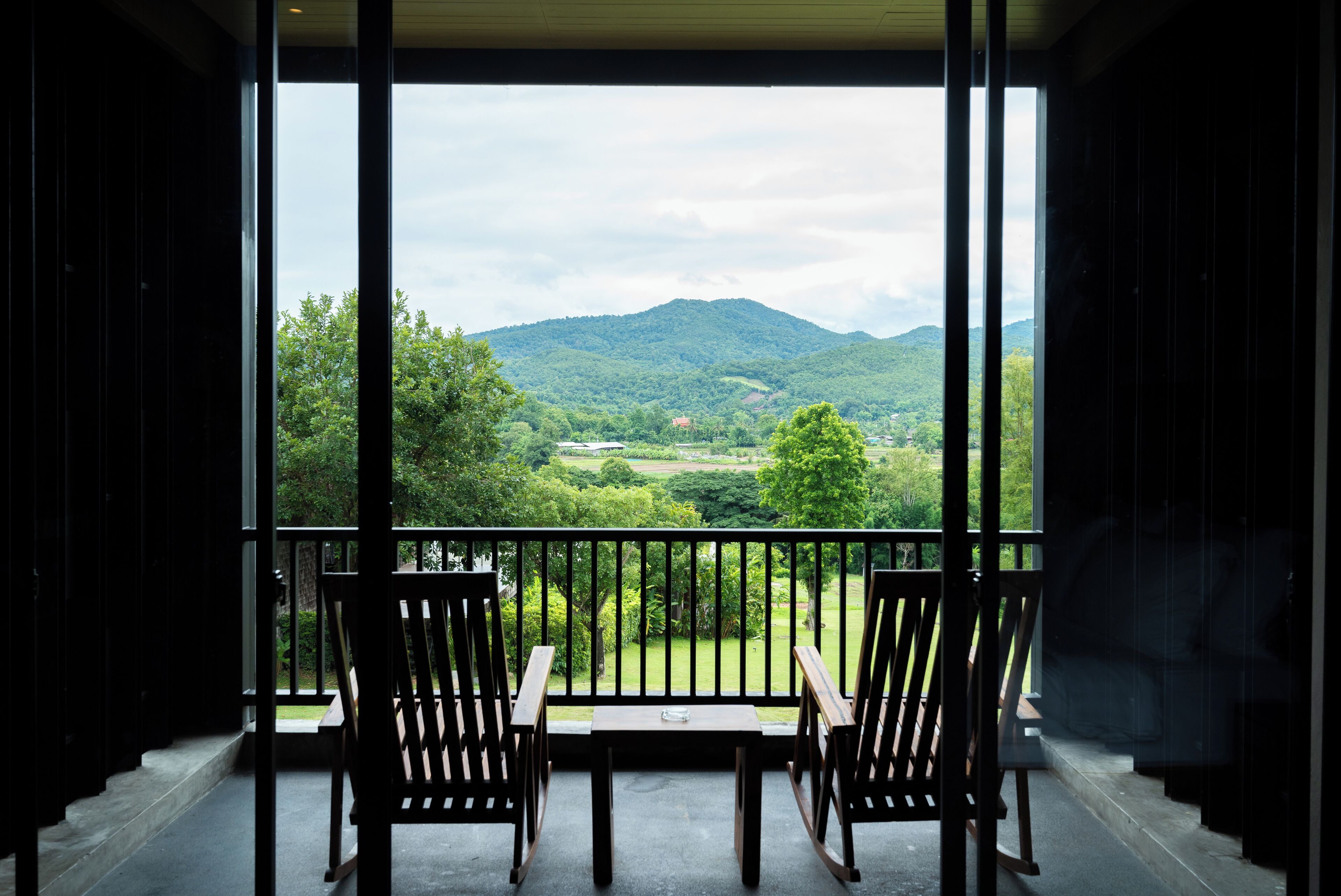Un balcón con dos sillas y una mesa de madera, con vista a un paisaje verde exuberante y montañas a lo lejos.