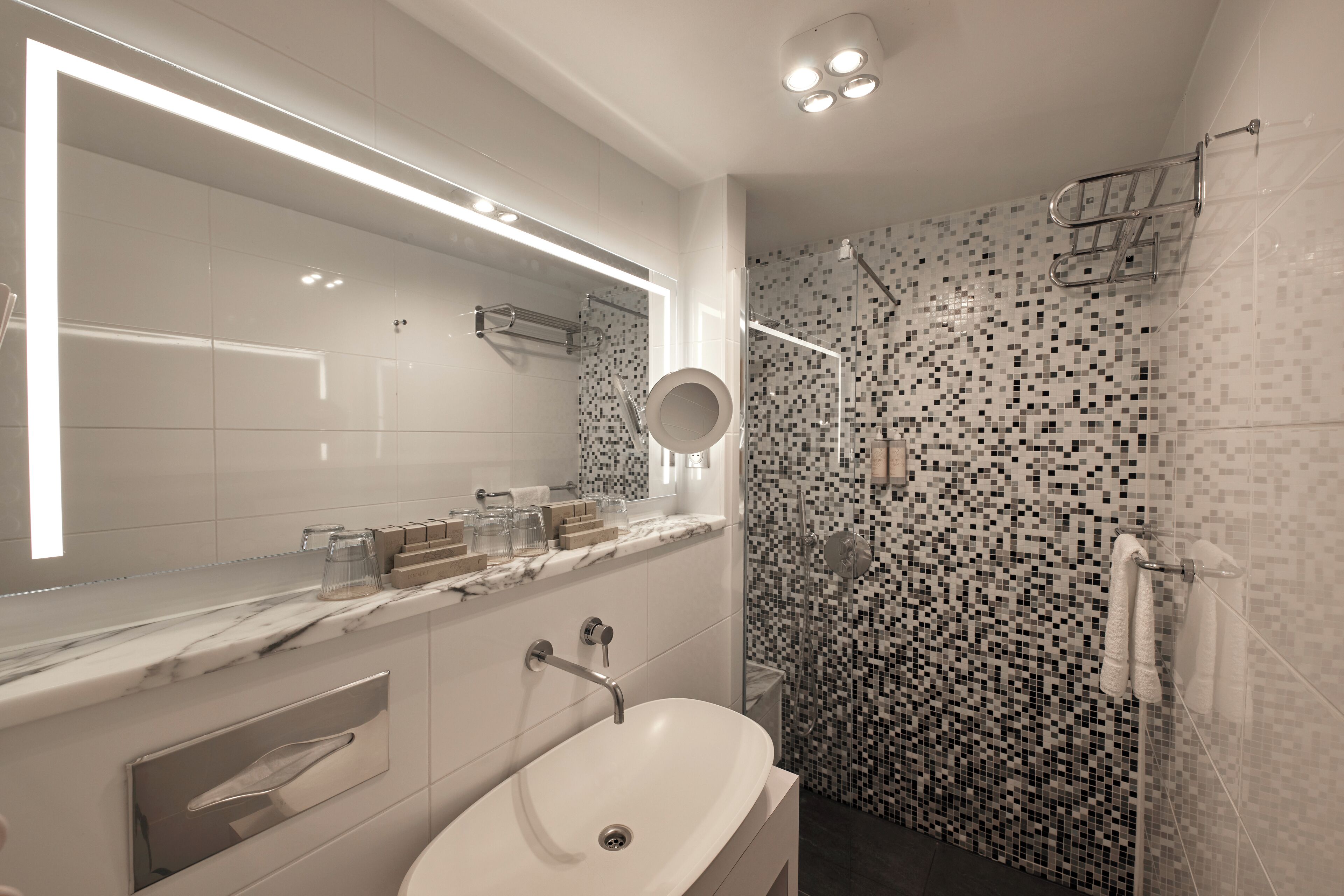 Un baño moderno con un espejo grande, encimera de mármol, lavabo blanco y ducha con mosaicos.