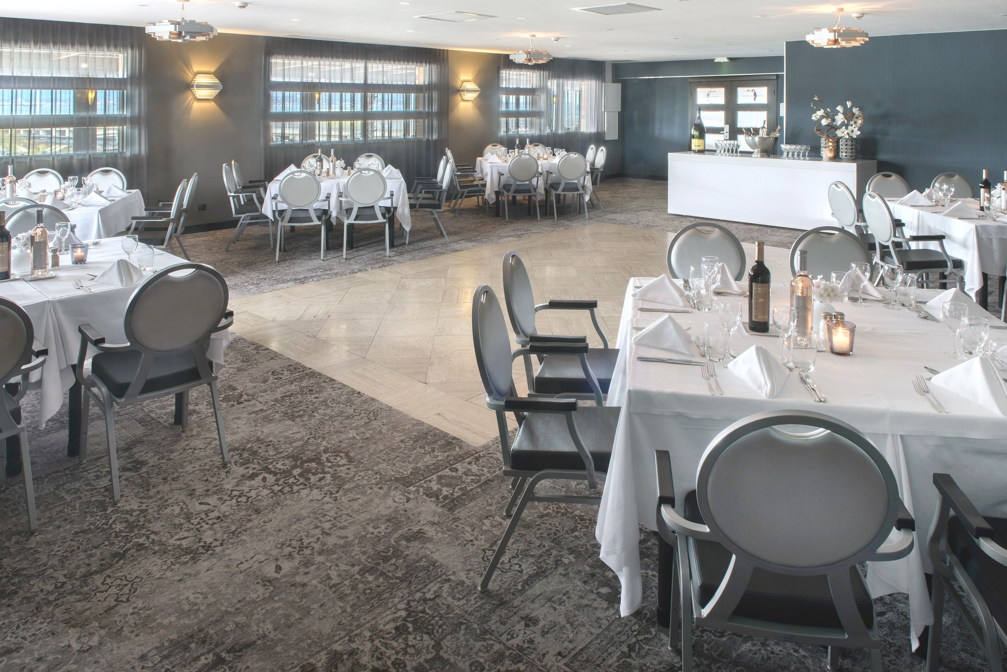 Un vaste espace dédié à la salle à manger, avec plusieurs tables dressées pour un événement formel, comprenant un bar central et de grandes fenêtres laissant entrer la lumière naturelle.