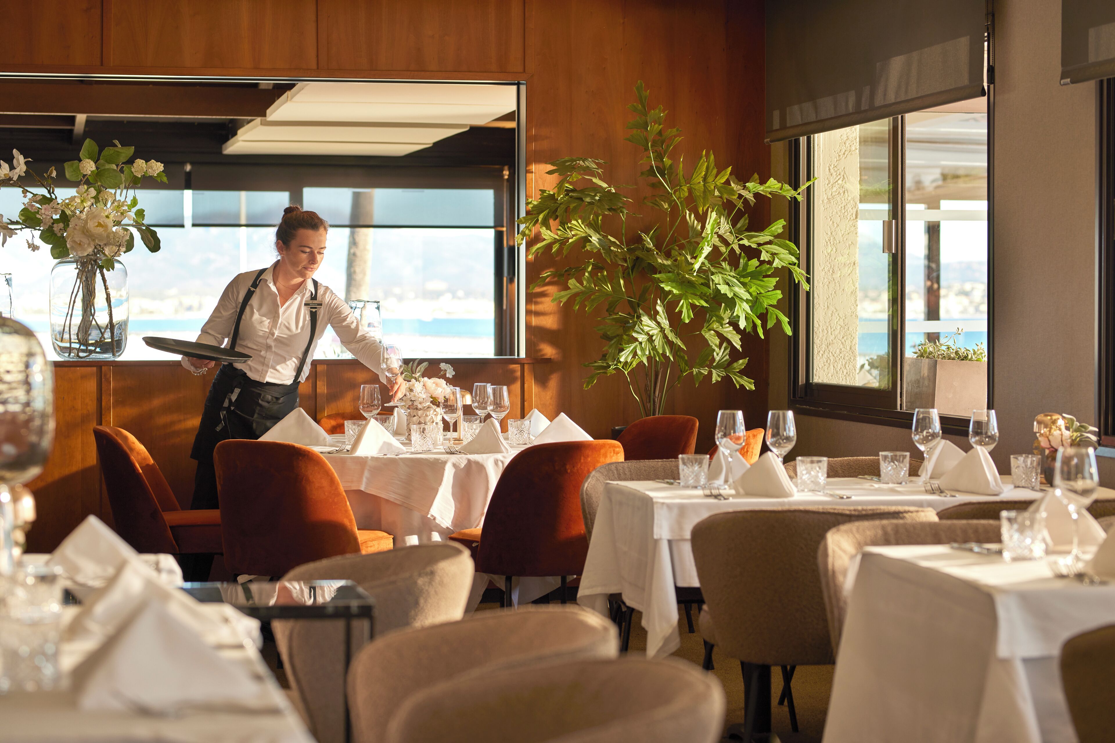 Un restaurant avec des tables dressées, un serveur en activité et une vue sur l’extérieur.