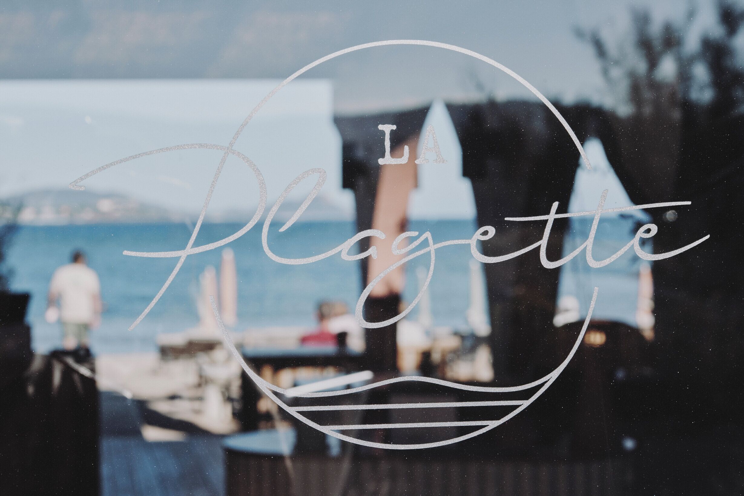 Ein Fenster mit dem Schriftzug „LA Plage“ und Blick auf das Meer und Boote.