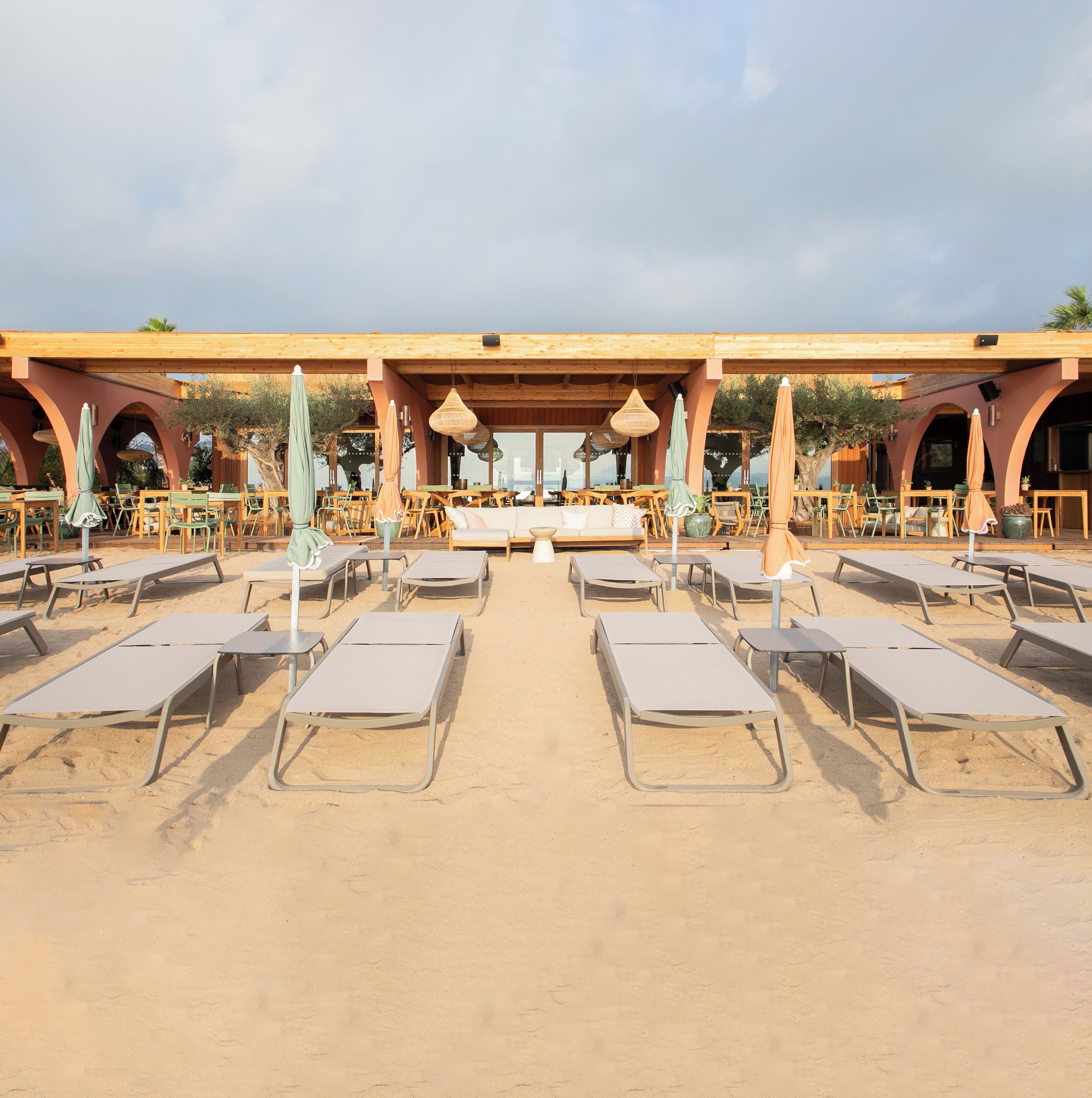 A beachside lounge area with sun loungers and tables under a wooden pergola, overlooking the sea.