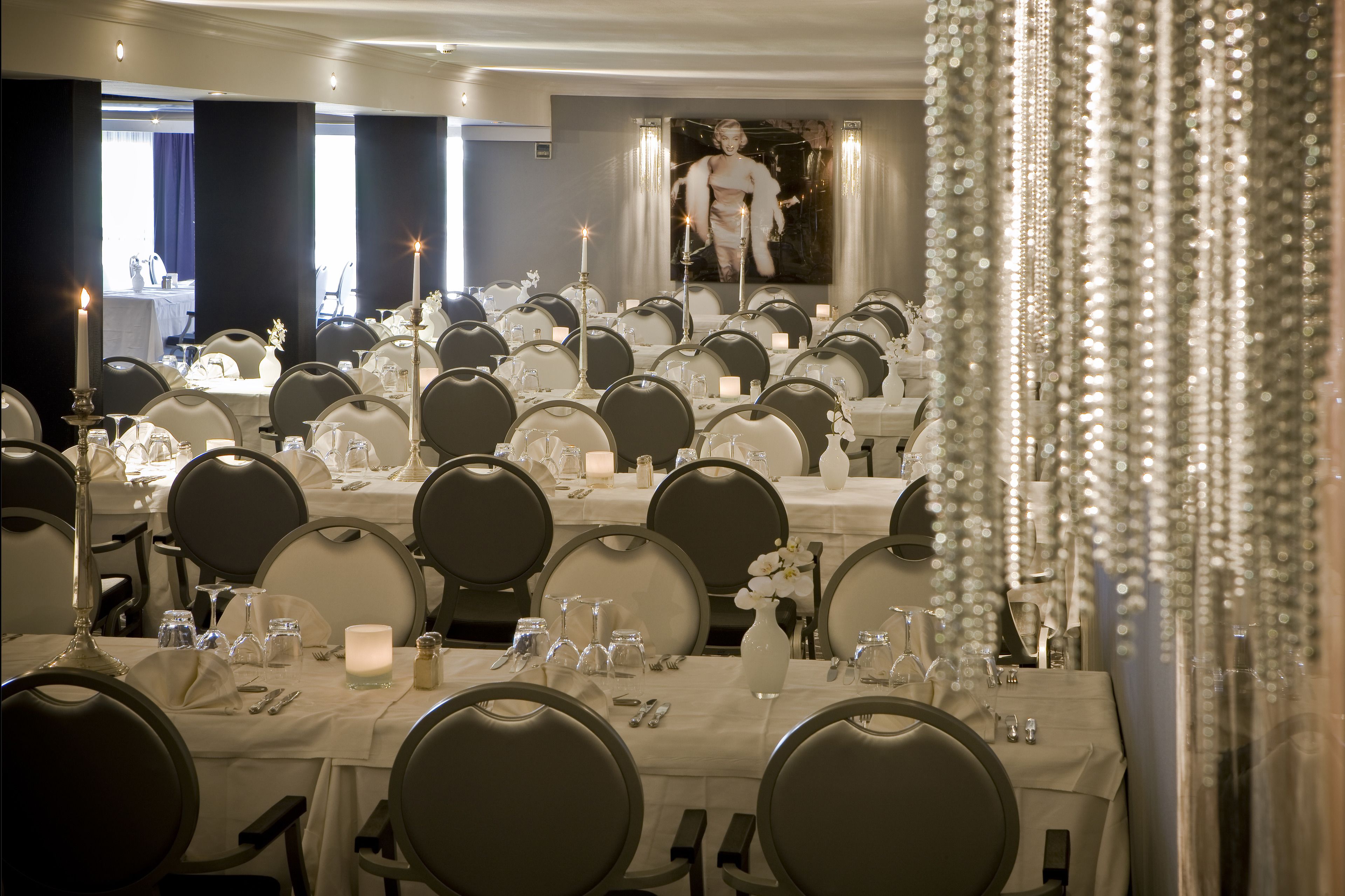 A formal dining room with multiple tables set for guests, featuring white tablecloths, candles, and elegant table settings.