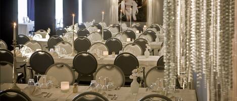 A formal dining room with multiple tables set for guests, featuring white tablecloths, candles, and elegant table settings.