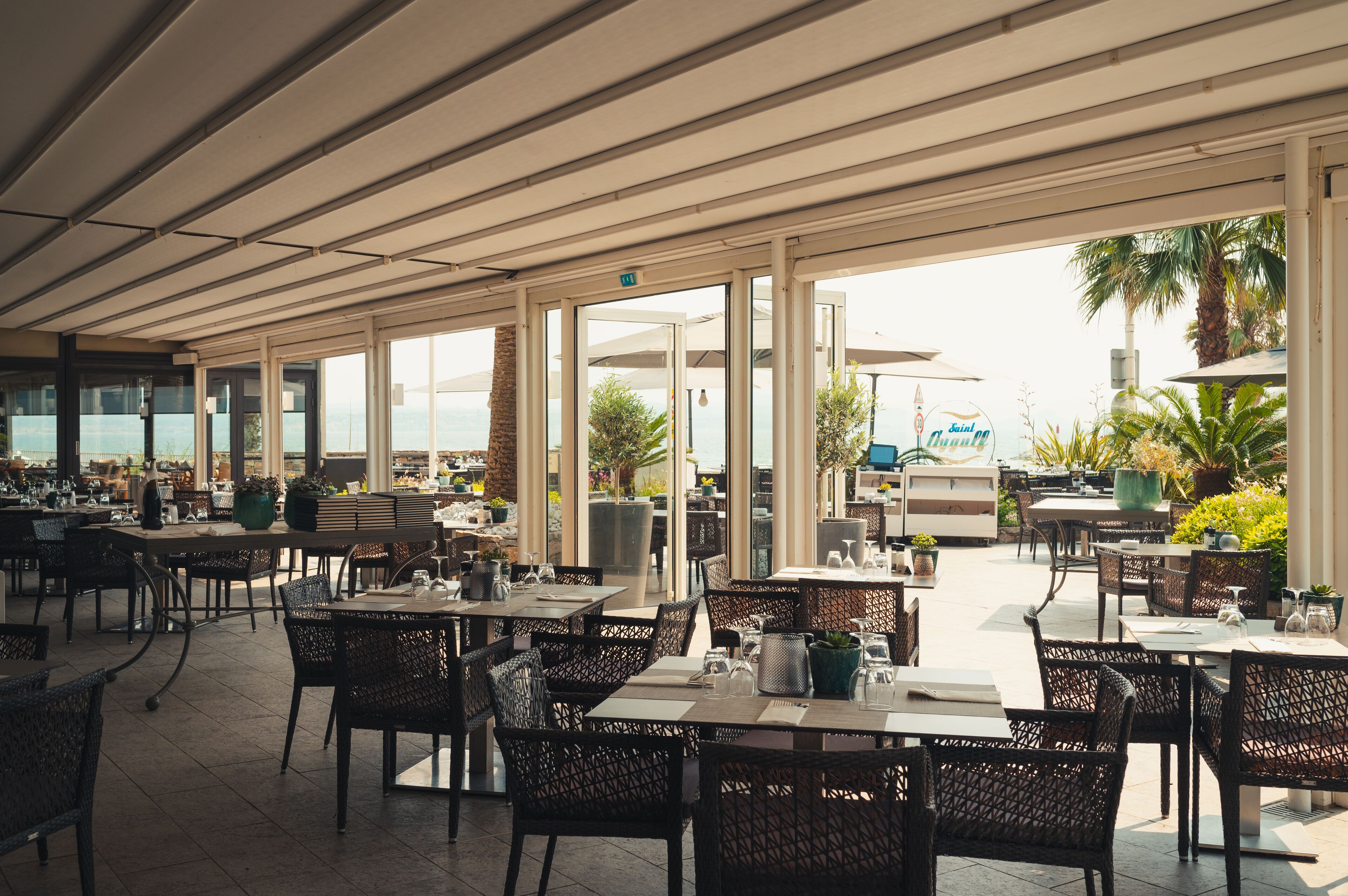 An outdoor dining area with tables and chairs, large windows, and potted plants.