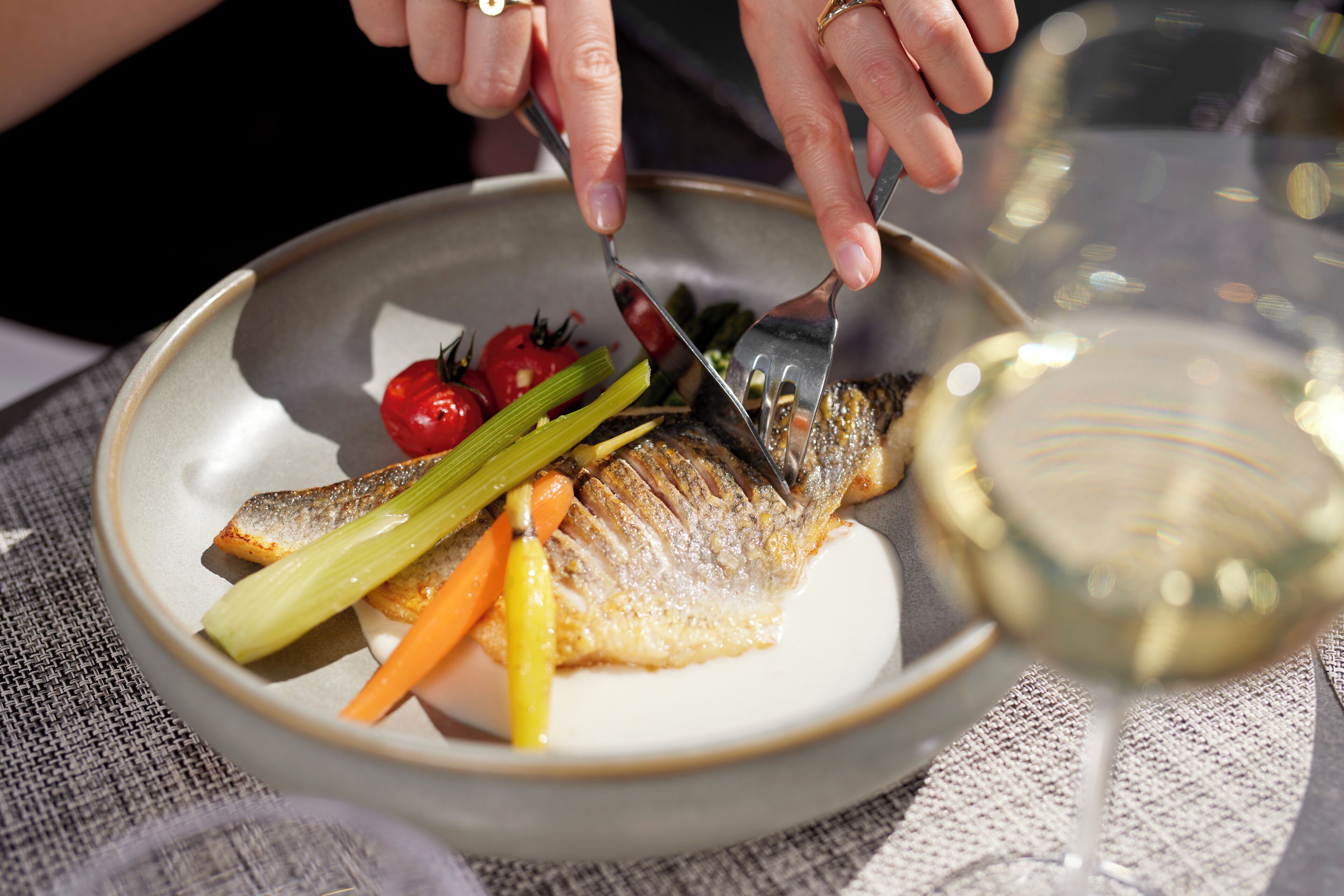Uma pessoa cortando um peixe em um prato com legumes e um copo de vinho branco.