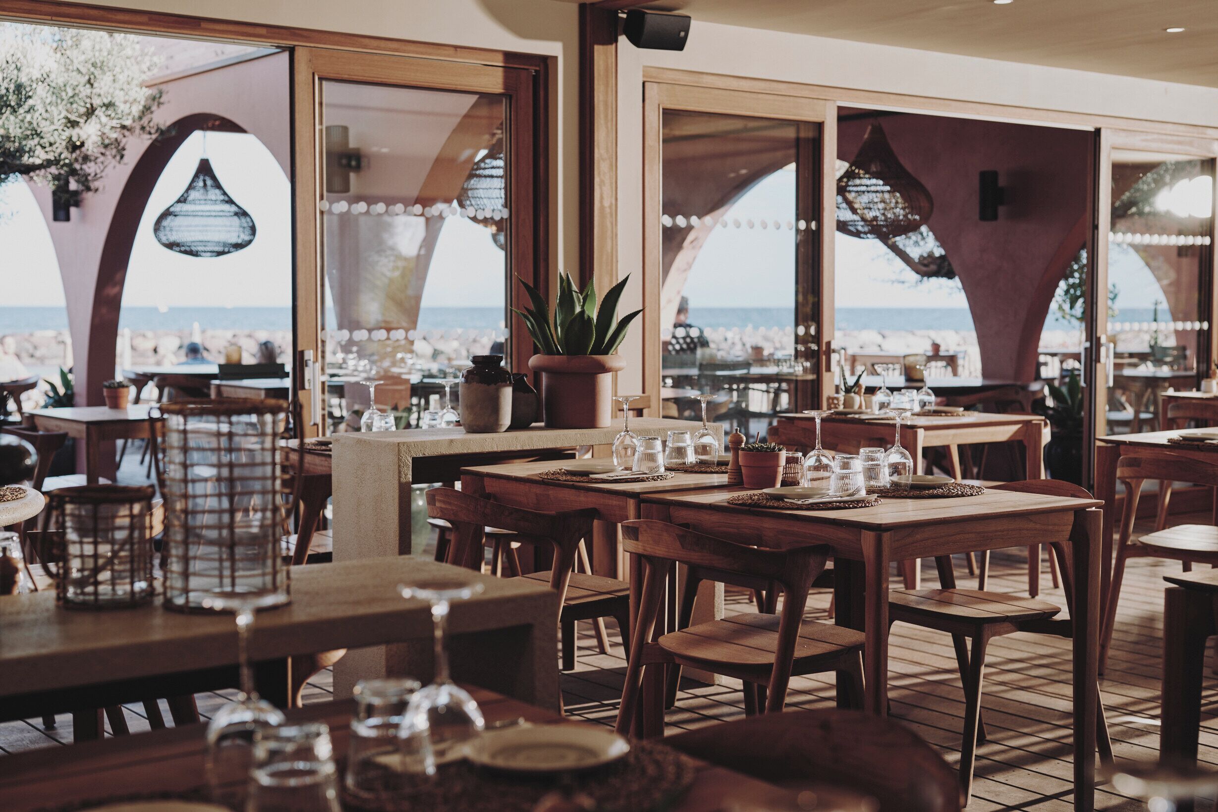 An outdoor dining area with wooden tables and chairs, potted plants, and a view of the beach.