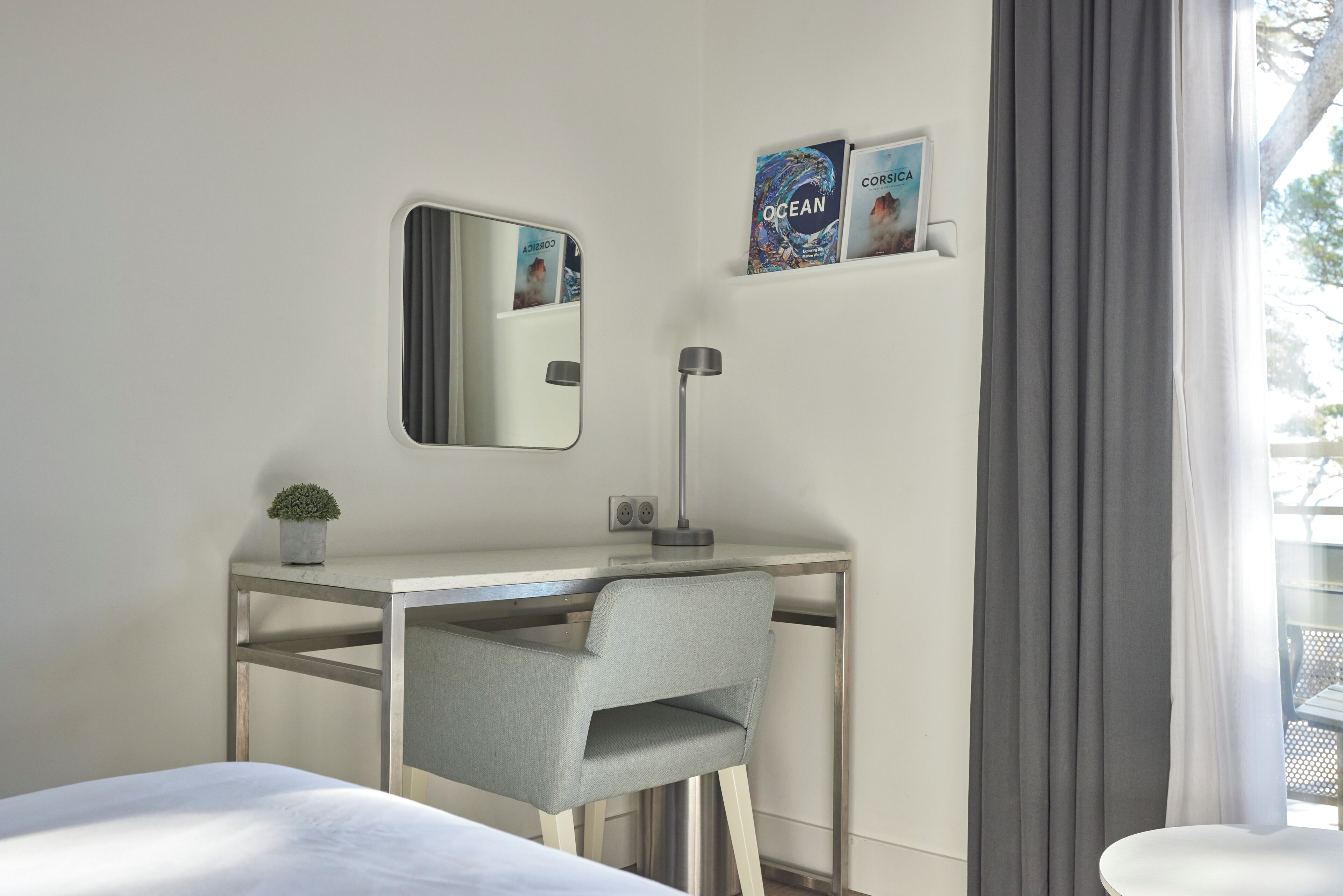 A hotel room with a bed, a desk with a chair, a mirror, a lamp, a small plant, and a shelf with books.