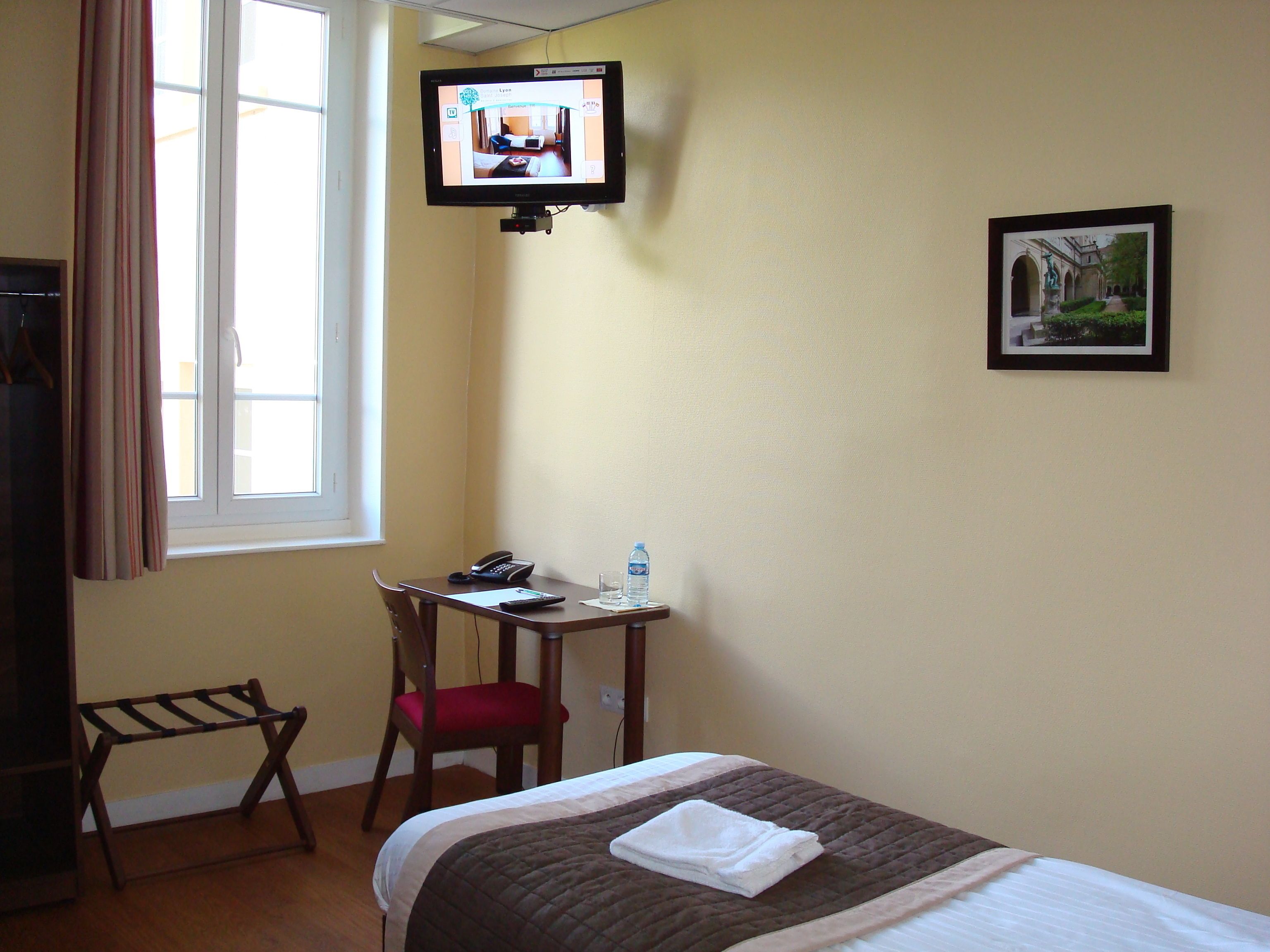 A hotel room with a bed, a desk with a chair, a television mounted on the wall, and a window with curtains.