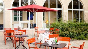 An outdoor patio with tables and chairs, a red umbrella, and a building with arched windows in the background. - Domaine Lyon Saint Joseph (Francheville)