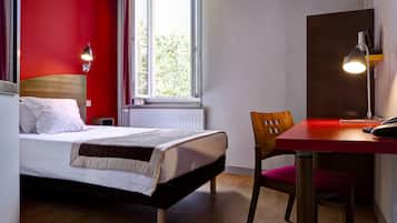 Une chambre d’hôtel avec un lit, un bureau avec une chaise, une lampe et une fenêtre avec des rideaux.