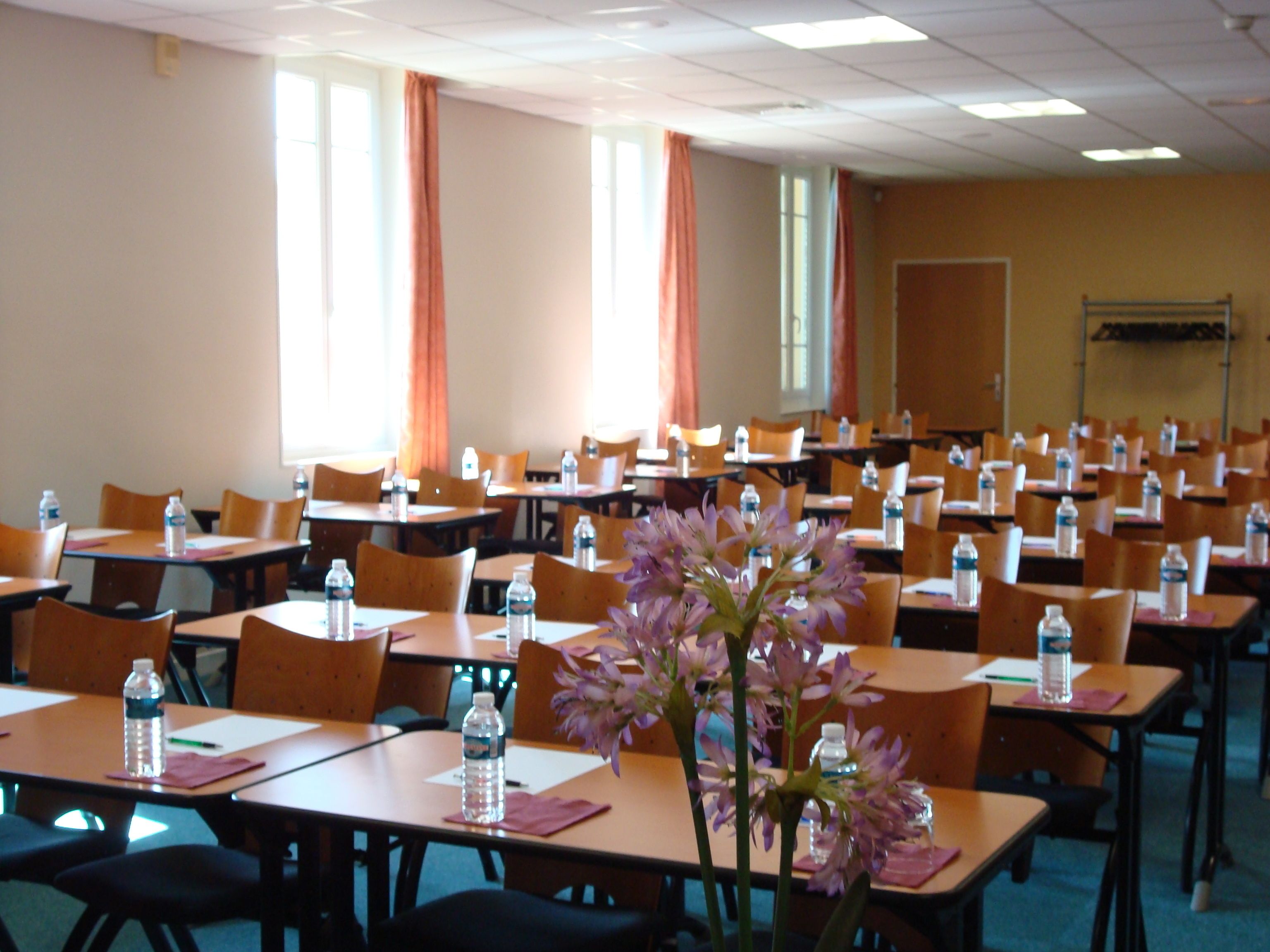 A conference room with rows of tables and chairs, water bottles on each table, and a large window with curtains.