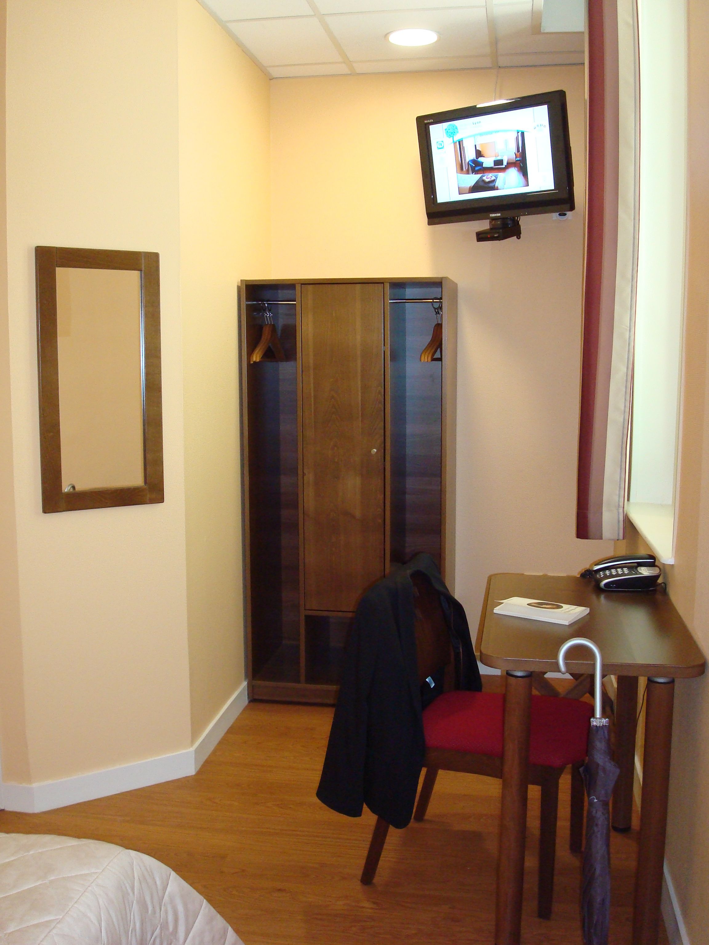 Een hotelkamer met een houten garderobe, een tafeltje met een telefoon en een afstandsbediening, een stoel en een aan de muur bevestigde televisie.