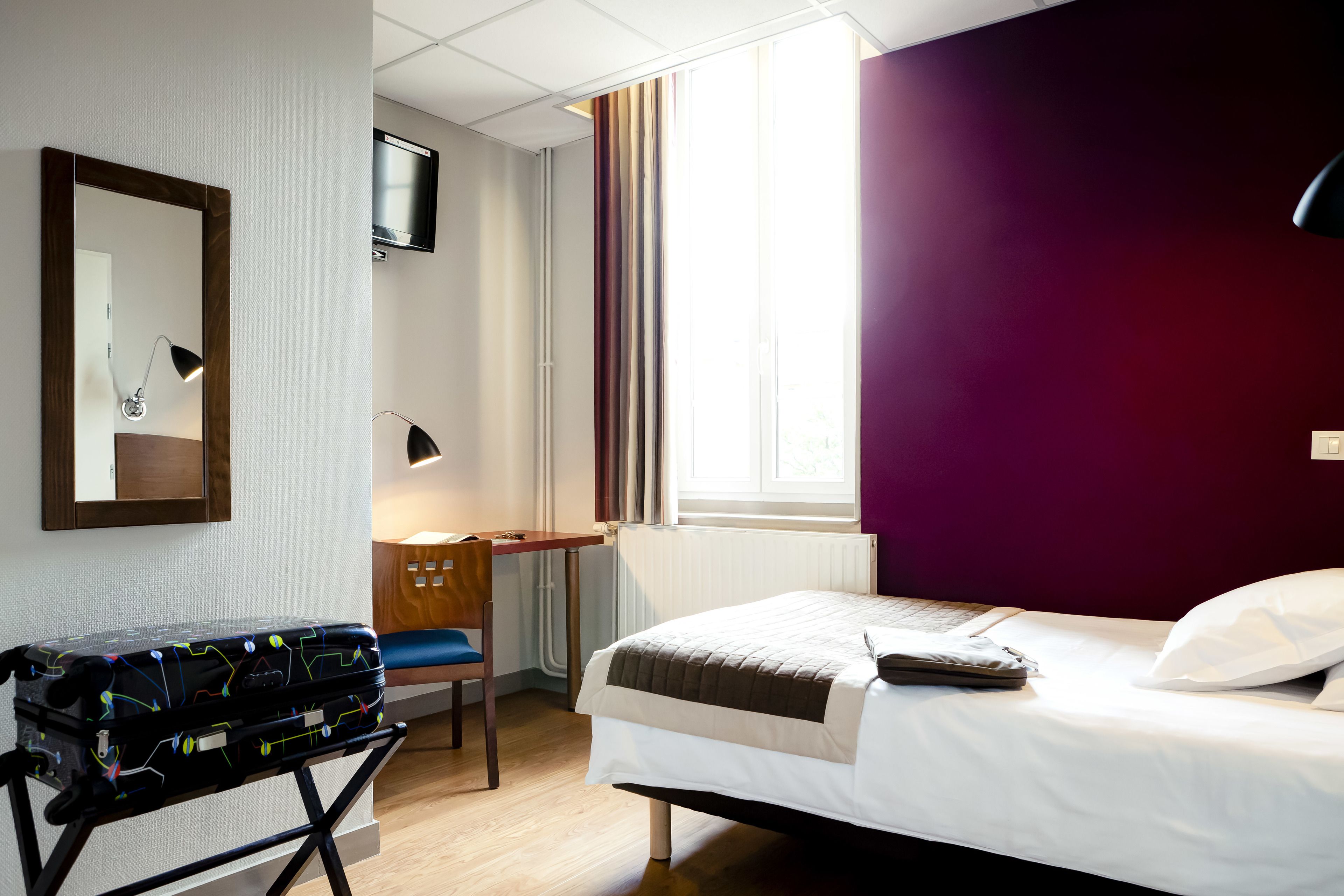 A hotel room with a bed, a desk, a chair, a suitcase, and a television on the wall.