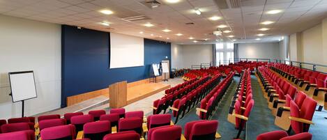 Un auditorio con hileras de sillas rojas, un atril, una pizarra y una pantalla de proyector.