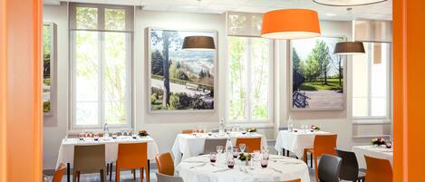 Un restaurante con mesas redondas, sillas anaranjadas y amplios ventanales con vistas al paisaje.