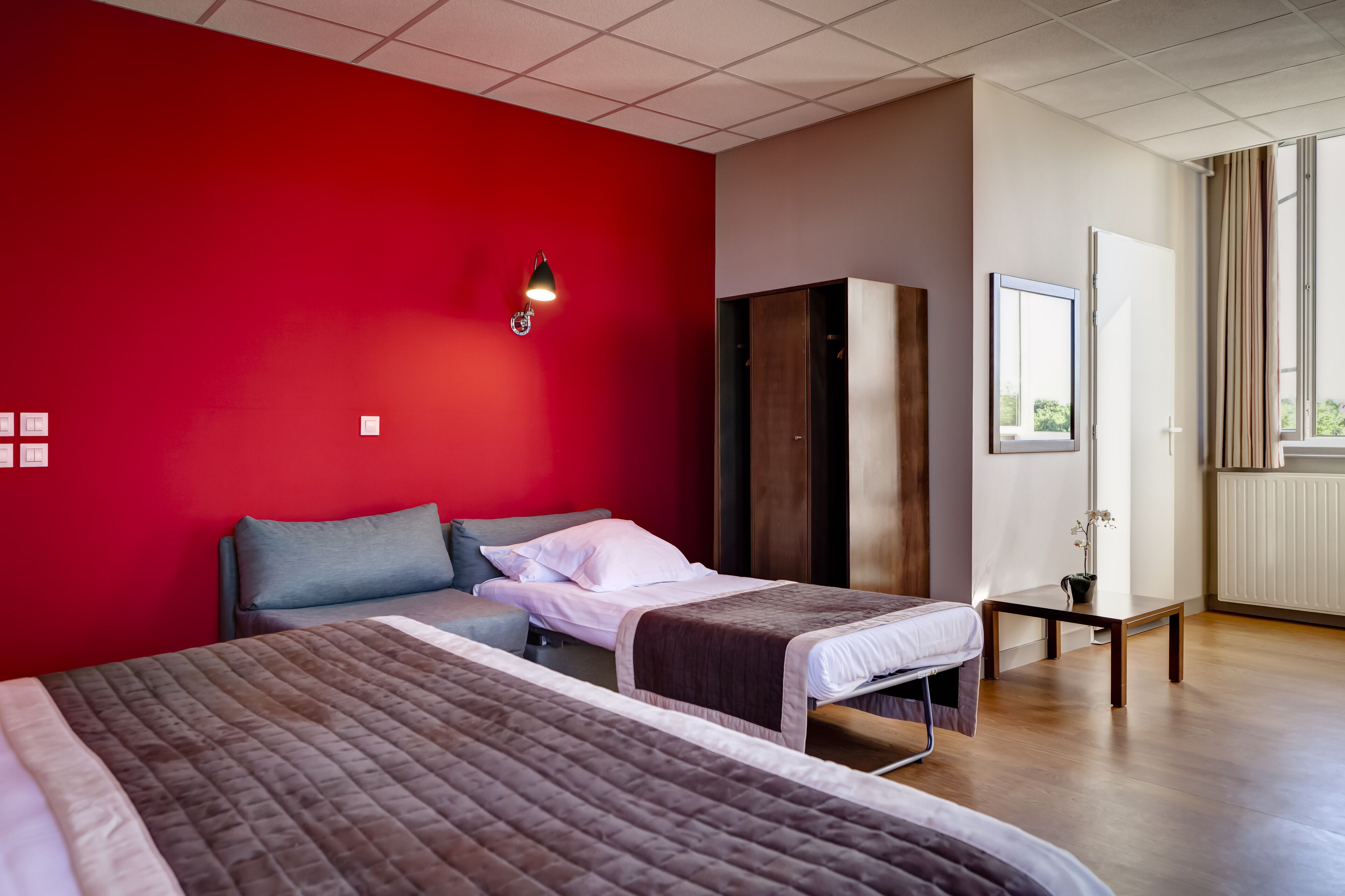 Habitación de hotel con dos camas, una pared roja, un armario de madera, una mesita y una ventana con cortinas.