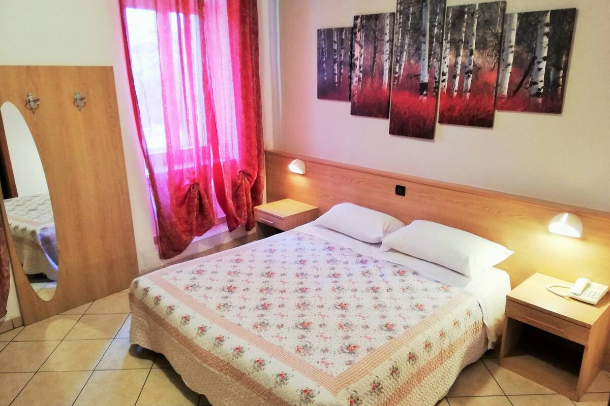 A hotel room with a bed, a nightstand, a mirror, a wooden headboard, and a wall with three paintings.