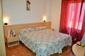 A bedroom with a wooden headboard, a floral bedspread, a nightstand with a bottle and remote, and a red curtain. - Hotel Neni (Brentonico)