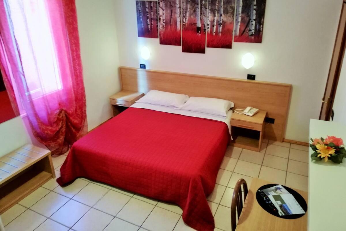 A hotel room with a large bed, a bedside table with a phone, a small desk with a chair, and a wall with abstract art.