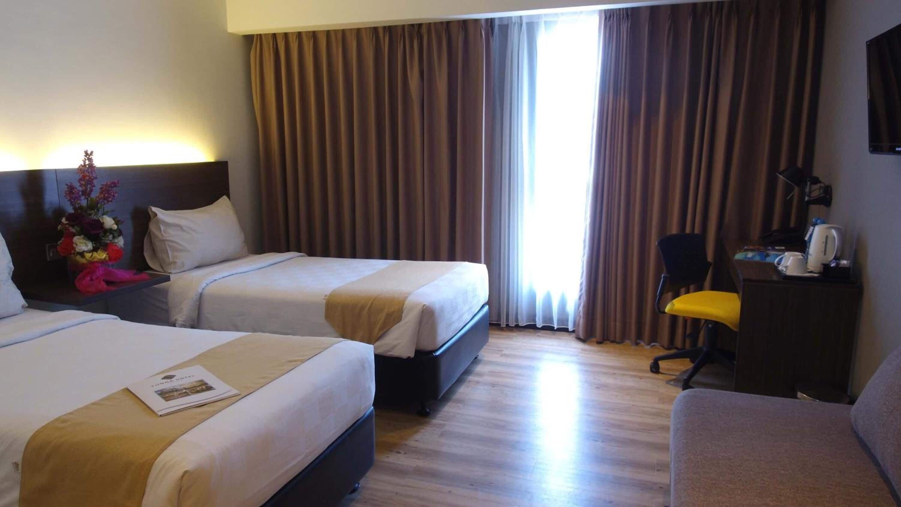 Ein Hotelzimmer mit zwei Betten, einem Schreibtisch mit Stuhl, einem Flachbildfernseher und einem großen Fenster mit Vorhängen.
